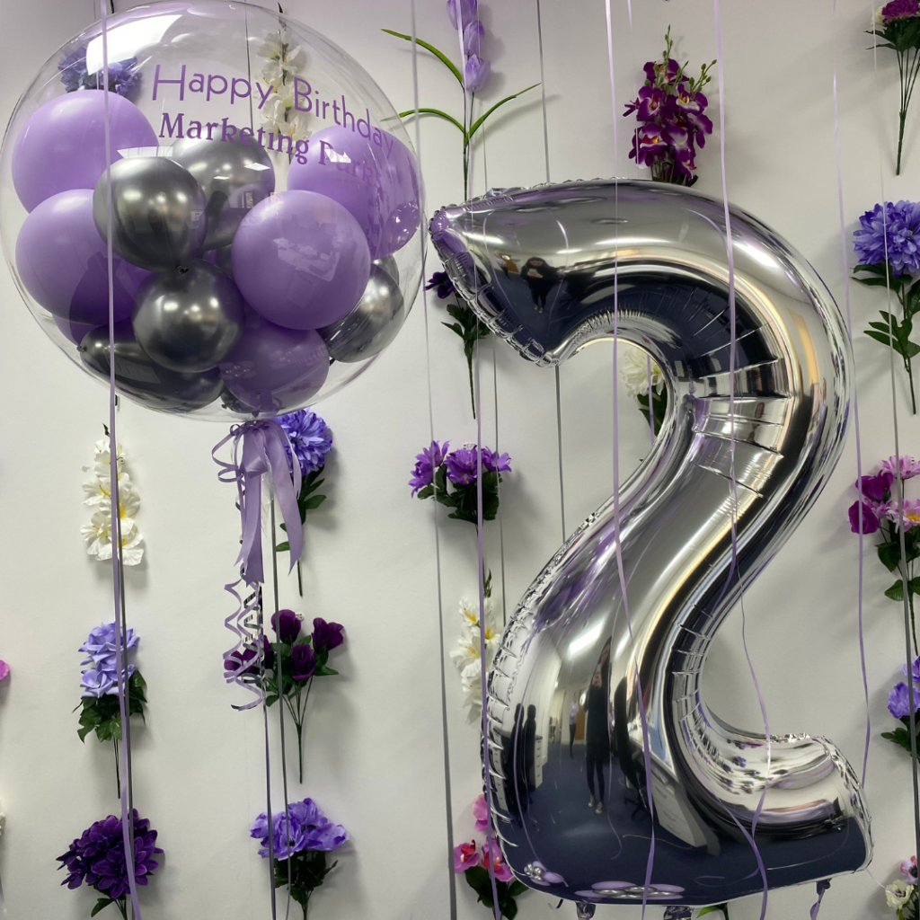 A photo of our office with a number 2 inflatable balloon. Behind is our flower wall which has lots of purple flowers, petals to reflect our branding.